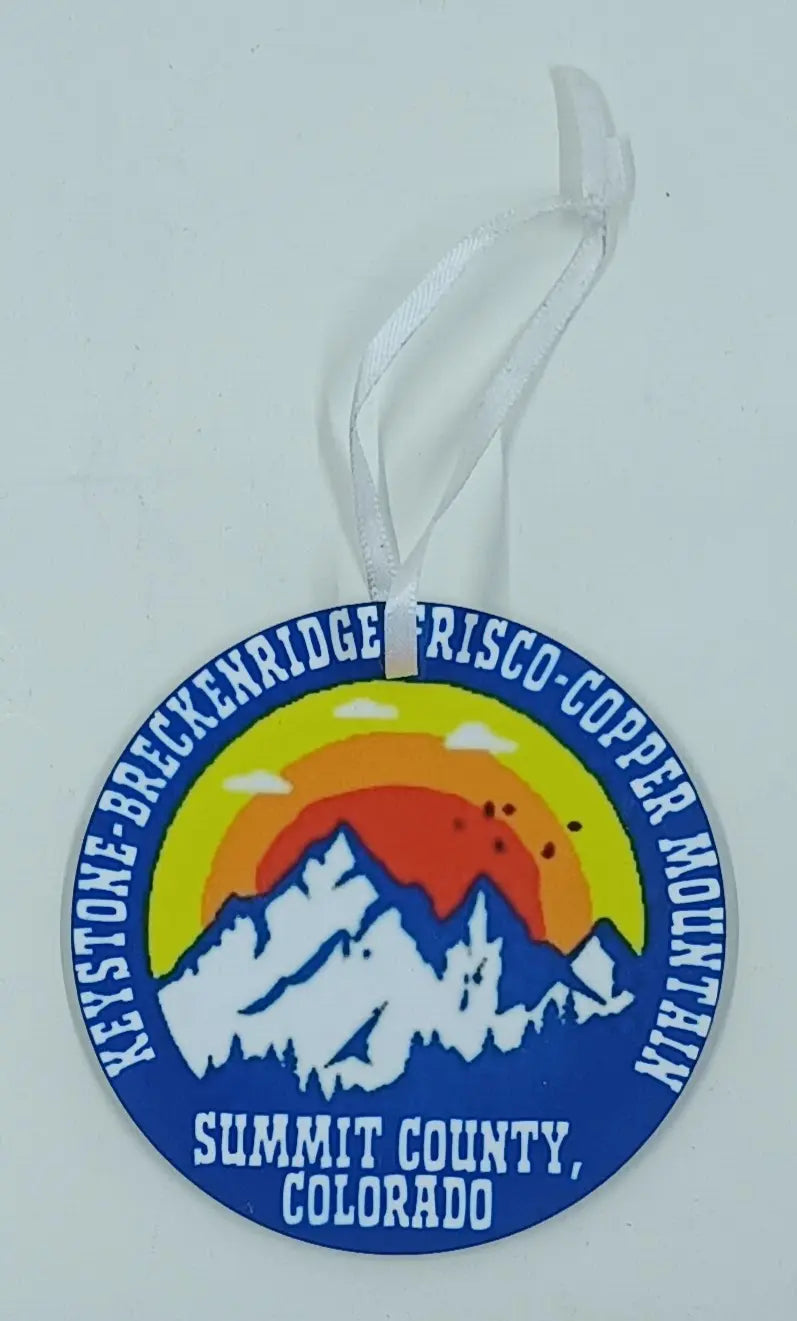 This is a round christmas ornament that has blue snow covered mountains and sunset behind it. It lists all the towns in summit county. 